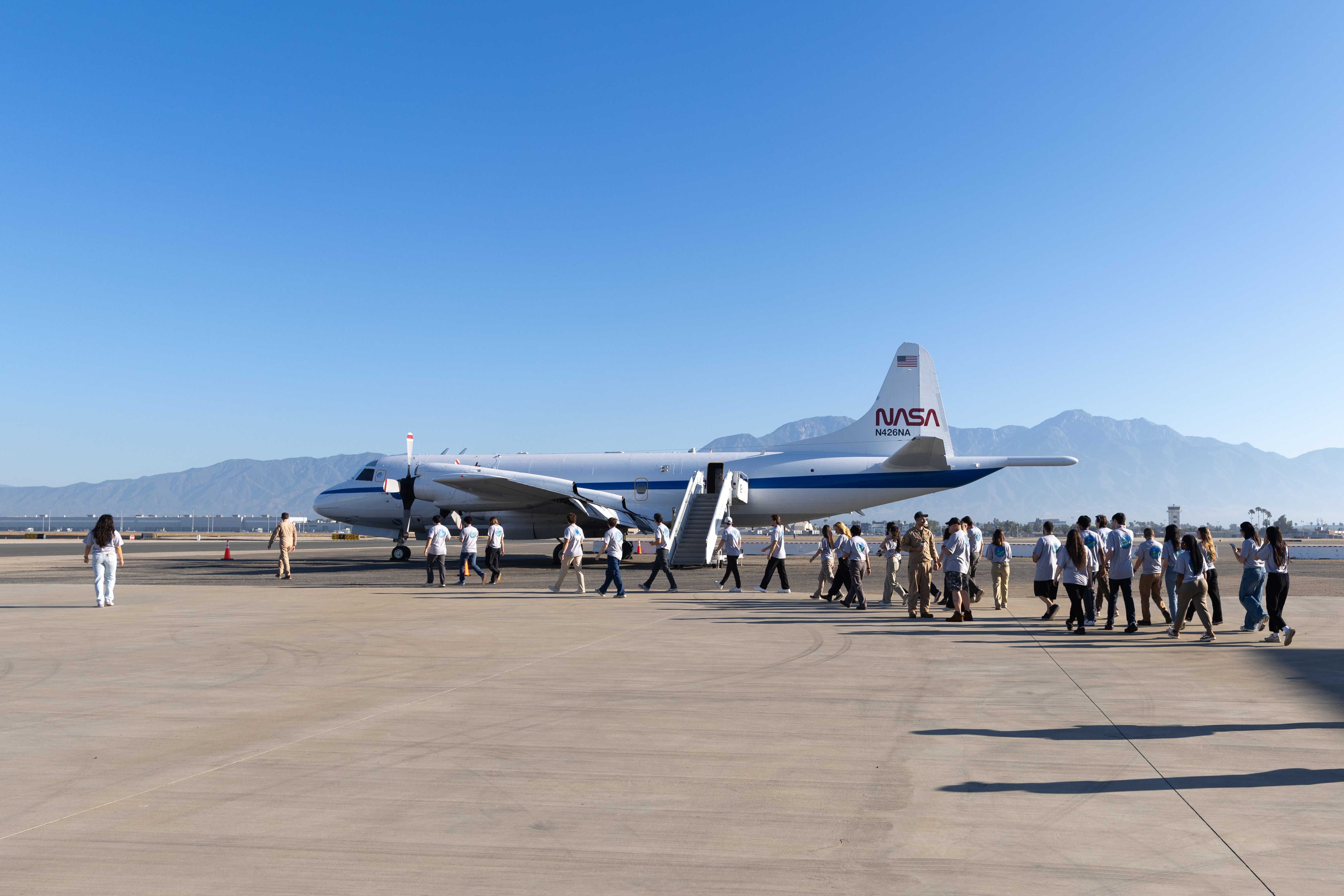 A line of students files across tan tarmac towards a large white aircraft with a blue stripe down the middle, with blue skies and a mountain in the background.