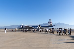 A line of students files across tan tarmac towards a large white aircraft with a blue stripe down the middle, with blue skies and a mountain in the background.