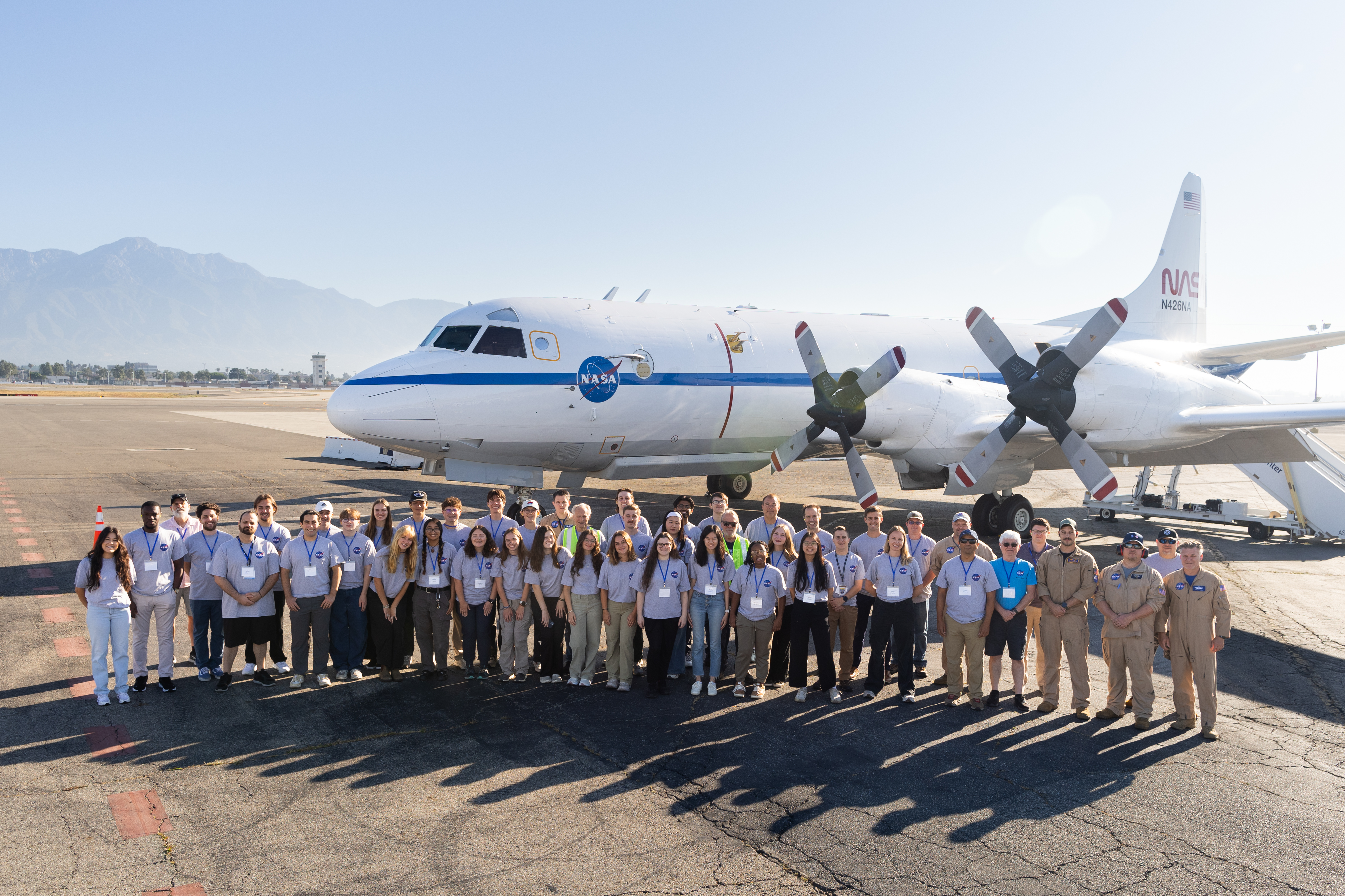 A group of students in gray shirts and several men in tan flight suits stand on tan tarmac in front of a large white plane with a blue stripe down the middle.
