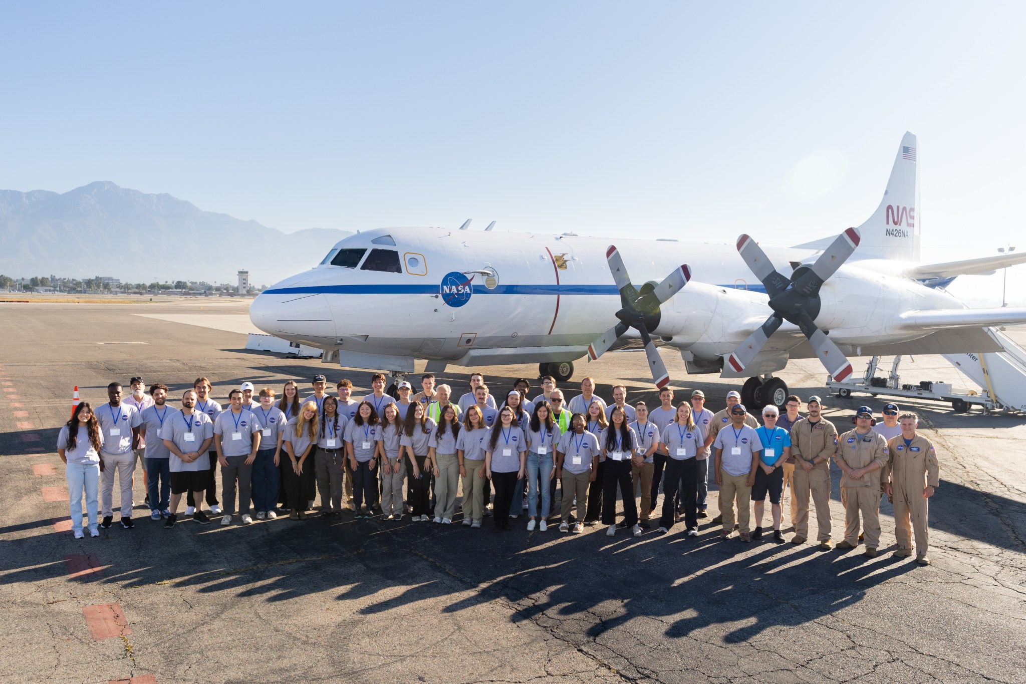 A group of students in gray shirts and several men in tan flight suits stand on tan tarmac in front of a large white plane with a blue stripe down the middle.