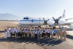 A group of students in gray shirts and several men in tan flight suits stand on tan tarmac in front of a large white plane with a blue stripe down the middle.
