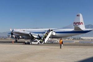 A line of students in yellow high-viz vests climbs a portable staircase to enter a large white aircraft with a blue stripe down the middle, with blue skies and a mountain in the background. On the tan tarmac in the foreground, a lone student wearing an orange high-viz vest stands with several bags.