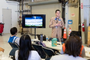 A man with short dark hair, wearing a tan flightsuit, stands in a hangar next to a portable screen with a graphic on it. Several students in the foreground sit with their backs to the camera.