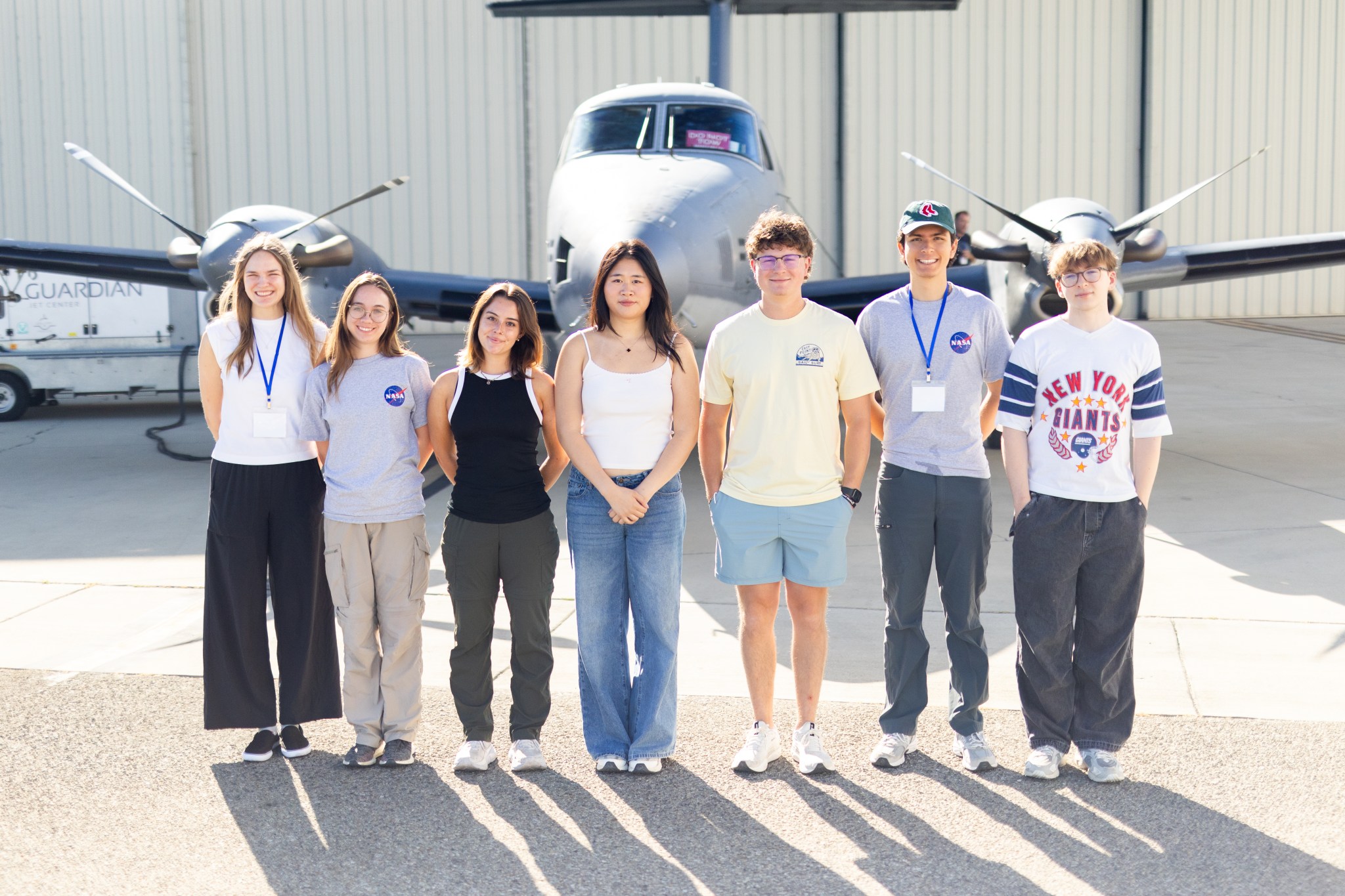 A group of seven students stand on tan tarmac in front of a small gray plane.