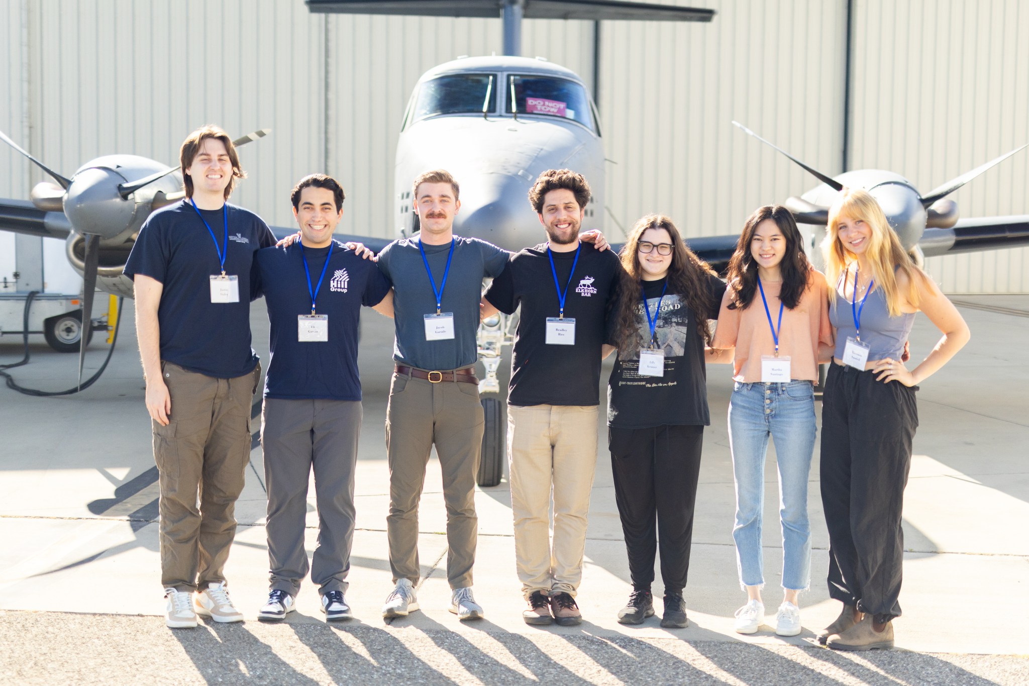 A group of seven students wearing badges on lanyards stand on tan tarmac in front of a small gray plane.