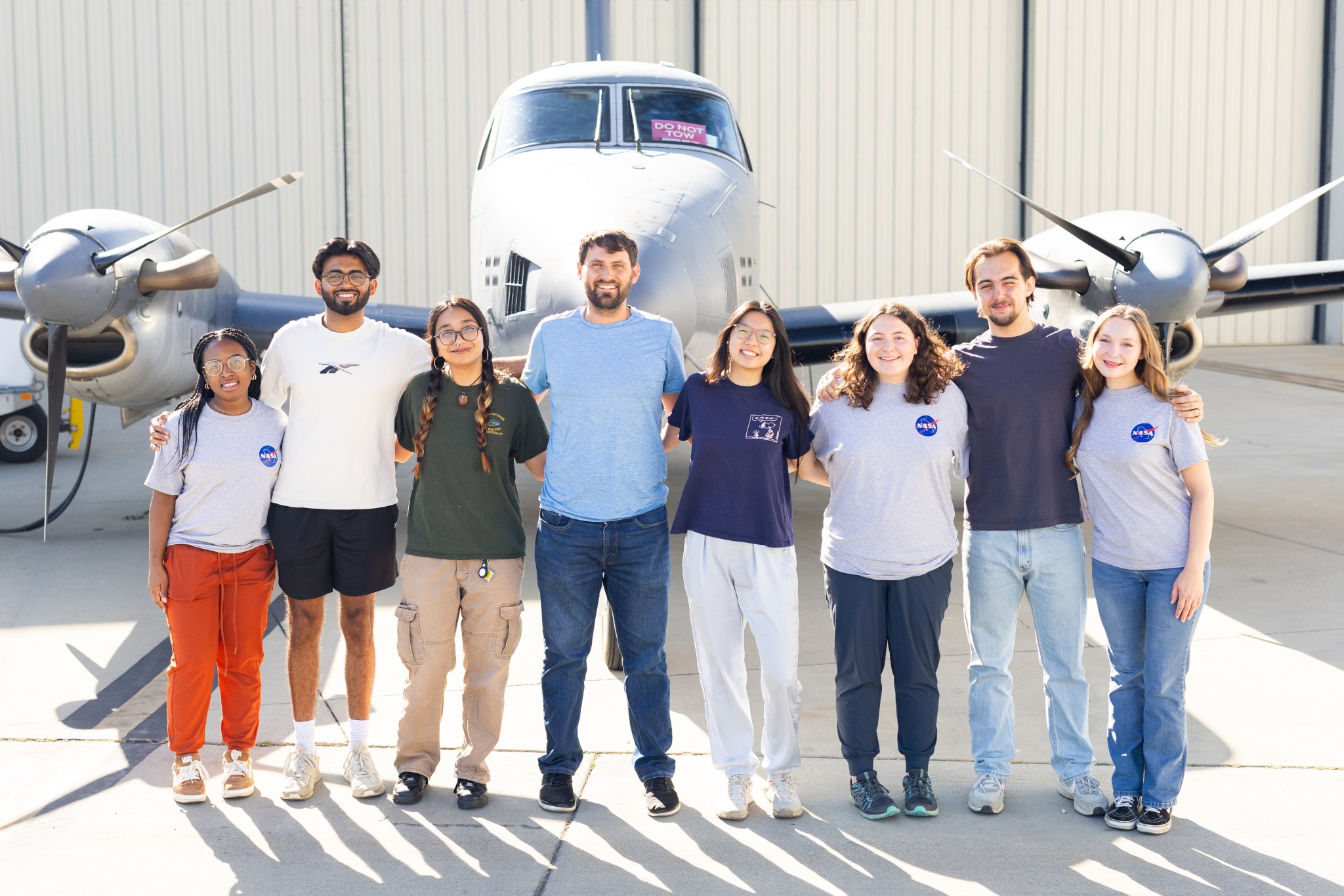 A group of eight people stand on tan tarmac in front of a small gray plane.