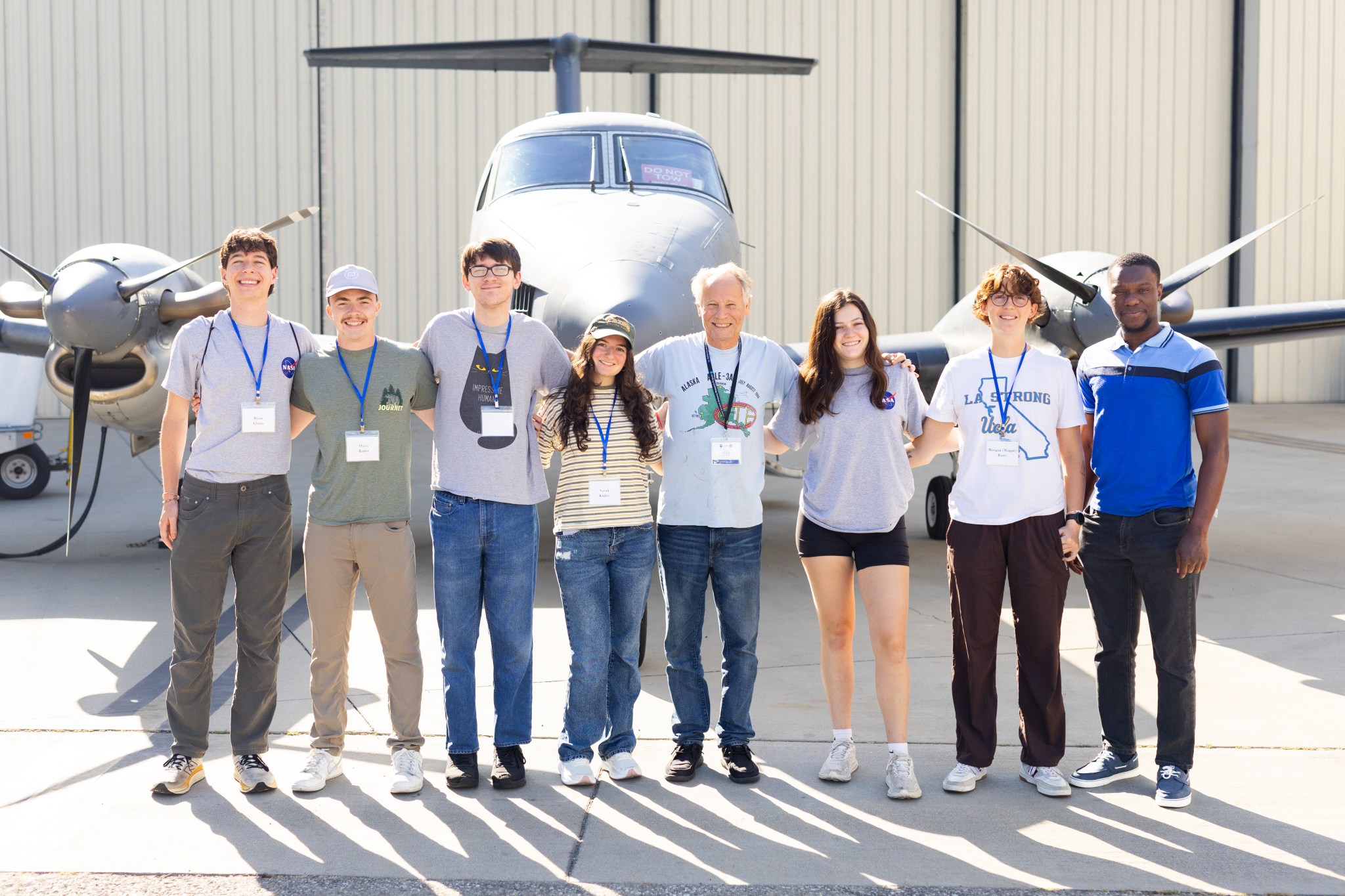 A group of eight people wearing badges on lanyards stand on tan tarmac in front of a small gray plane.