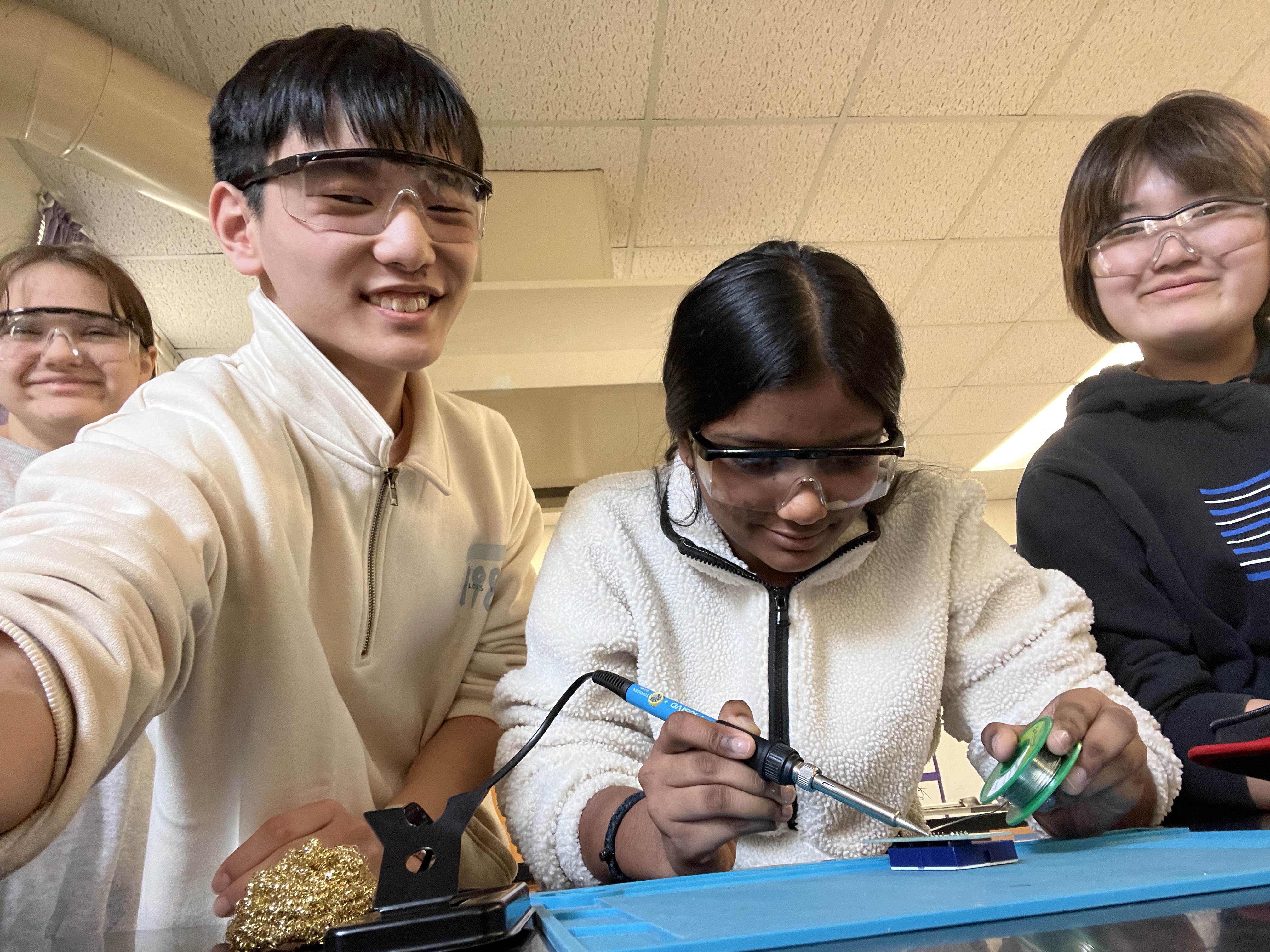 Three students smile for the camera as one student solders a small device. 