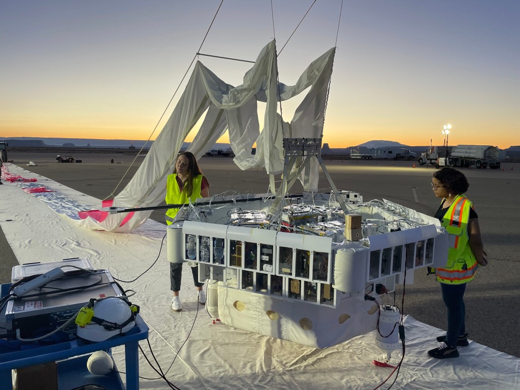 Two people in reflective vests make adjustments to a large gondola housing individual experiment containers and attached to a yet-to-be-inflated high altitude balloon at sunrise.
