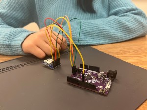 A student's hand is poised behind a device with colorful wires extending from one box to another.