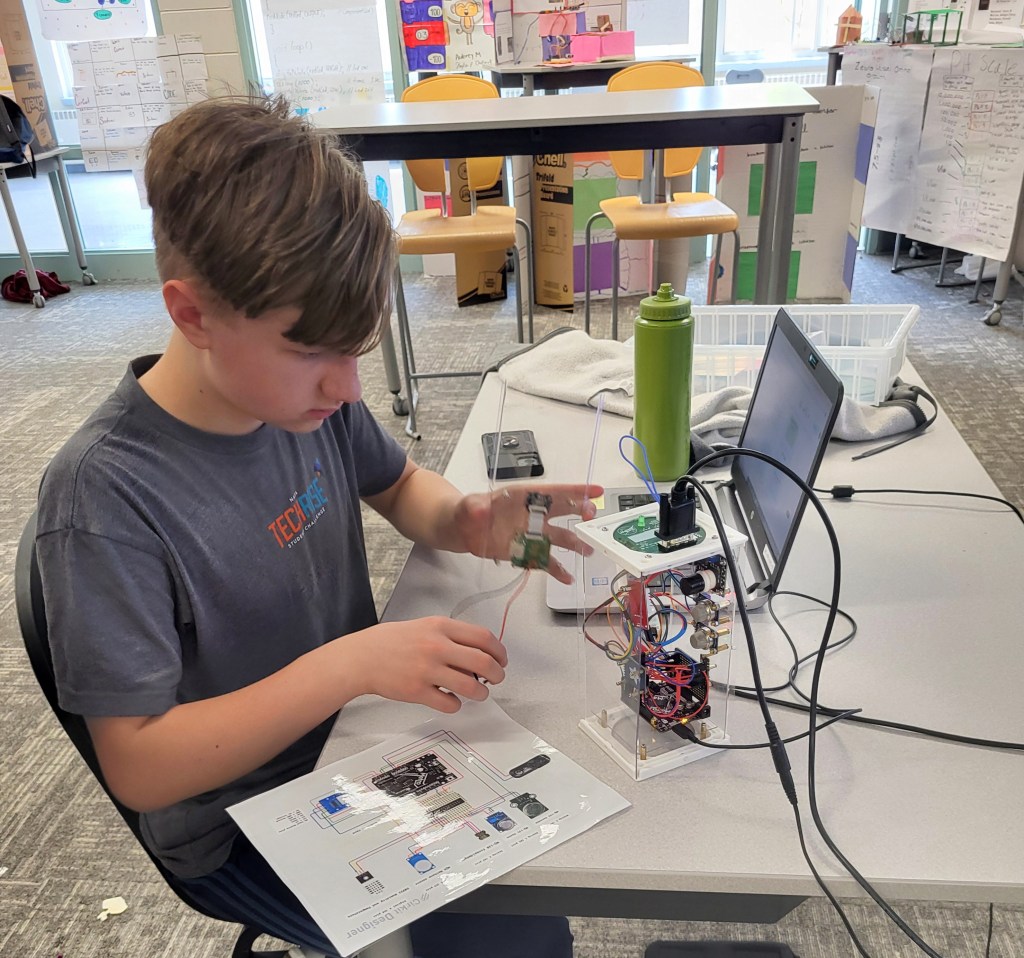 A student holds a device while looking at a clear rectangular container. A schematic drawing is on the table.