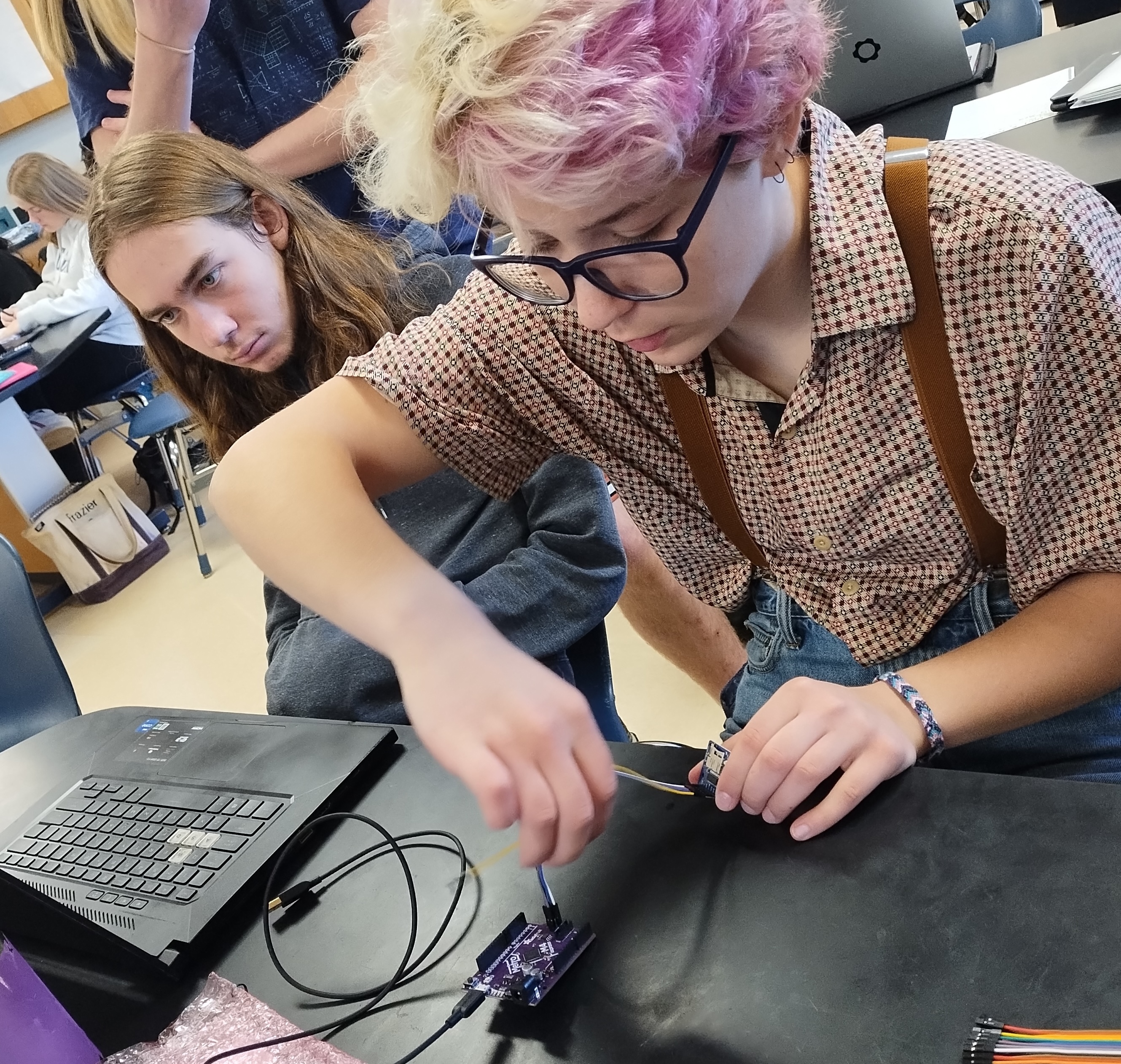 A student assembles a small device at a workstation as another student looks at a laptop screen.