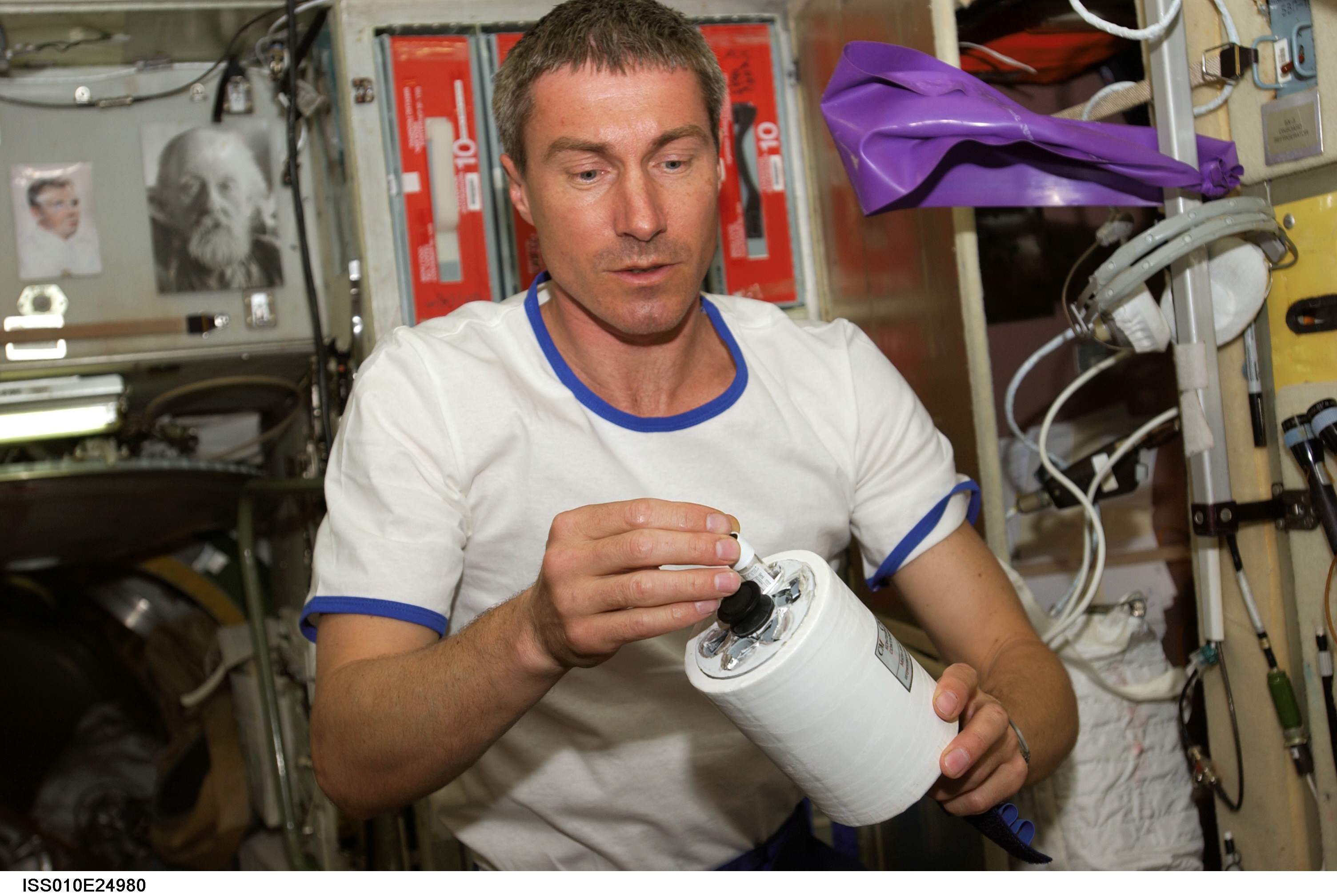 A crew member is seen working on the International Space Station. He is holding a small white cylinder with parts of the experiment inside of it.