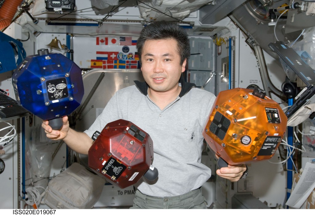 A space station crew member poses with three bowling-ball sized objects. One is blue, one is red, and one is yellow.