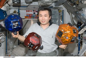A space station crew member poses with three bowling-ball sized objects. One is blue, one is red, and one is yellow.