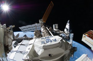The Alpha Magnetic Spectrometer experiment is seen installed on the external truss of the space station. The letters "AMS" appear on the experiment hardware. Space station radiators and solar arrays are in the background. The Space Shuttle is seen docked to the space station.
