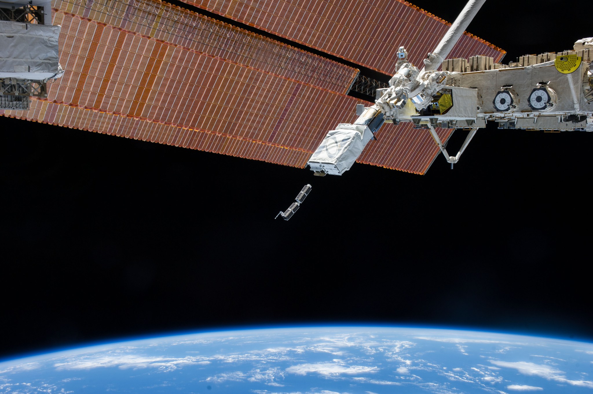 Small cubesats are seen coming out of the NanoRacks cubesat deployer. An orange colored space station solar array is behind the deployer.