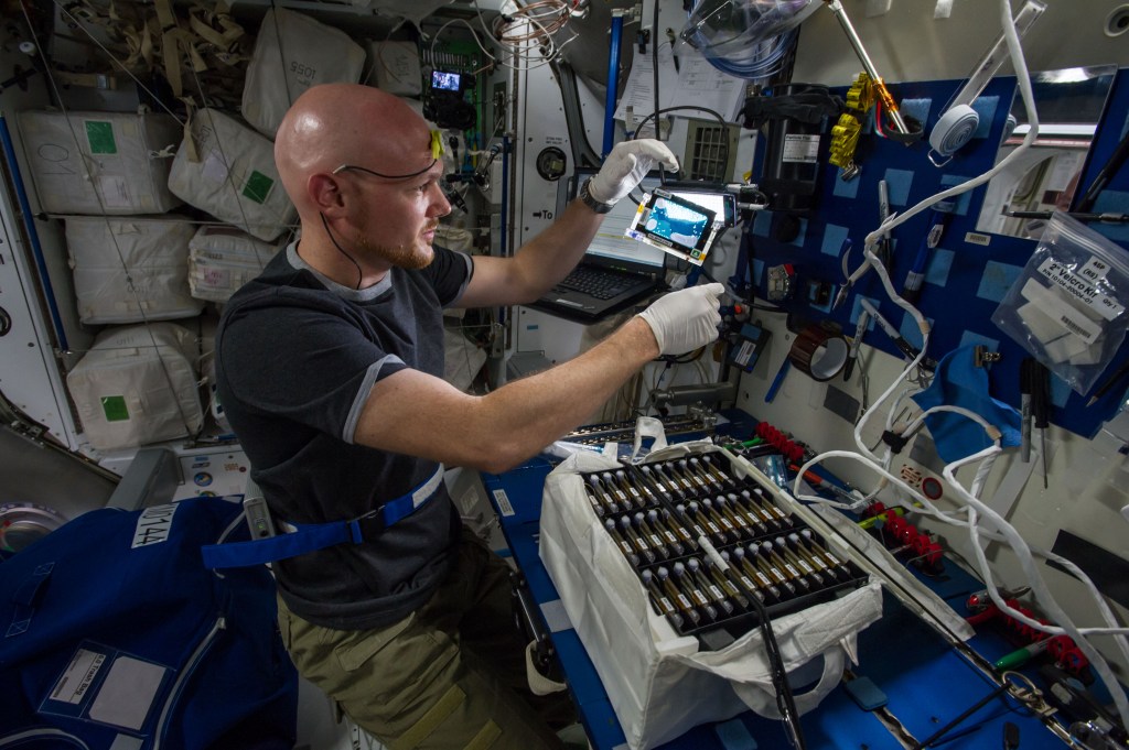 Astronaut Alexander Gerst is seen holding equipment for a space station experiment. There is a bag on a table next to Alexander. The bag is filled with additional payload hardware.