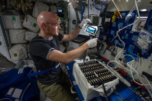 Astronaut Alexander Gerst is seen holding equipment for a space station experiment. There is a bag on a table next to Alexander. The bag is filled with additional payload hardware.