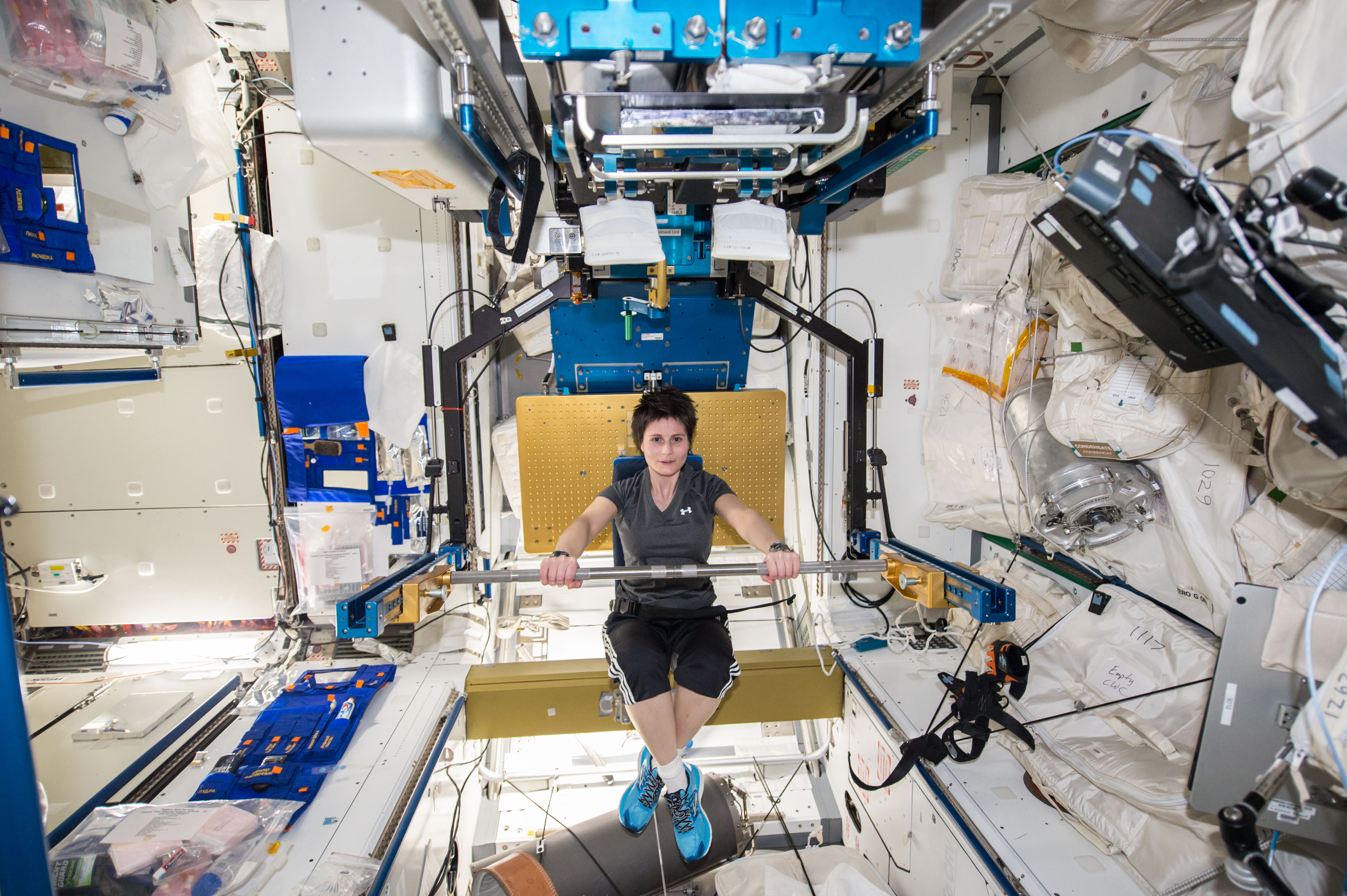 Astronaut Samantha Cristoforetti is seen performing exercises on the space station. Samantha is laying down on a yellow platform, and is pushing a bar away from her body.