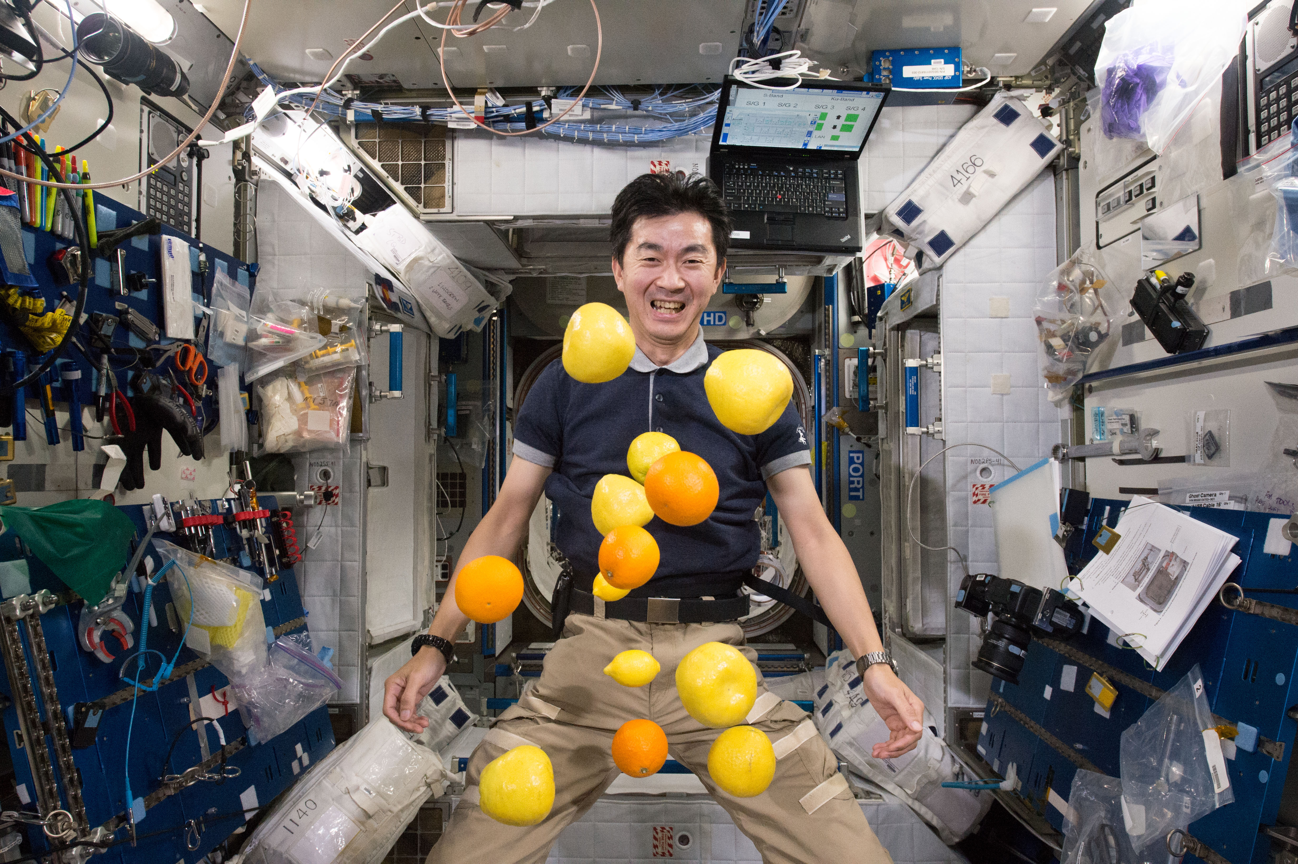 Astronaut Kimiya Yui is seen floating in the International Space Station. Numerous oranges and yellow colored apples and lemons float in front of Kimiya. Kimiya is smiling.