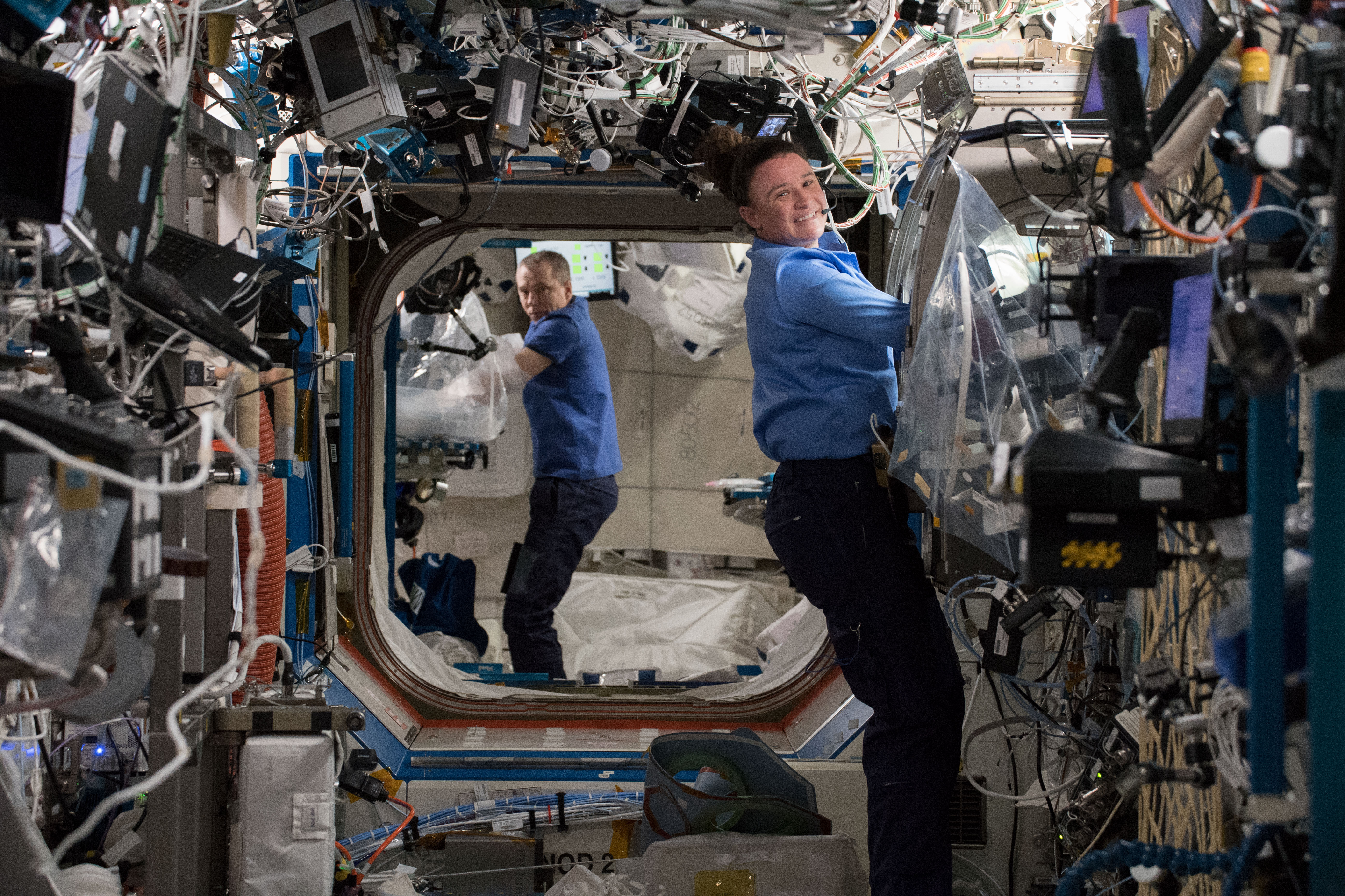 Astronauts Serena Auñón-Chancellor and Drew Feustel are seen conducting experiments using research gloveboxes on the space station. Serena is in the foreground on the right side of the image, and Drew is in the background on the left side. They are looking at the camera and smiling.