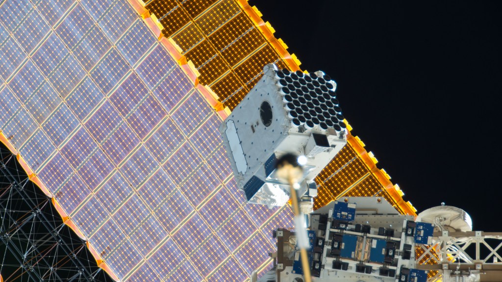 The NICER payload is seen installed at an exterior location on the space station. A space station solar array is seen in the background.