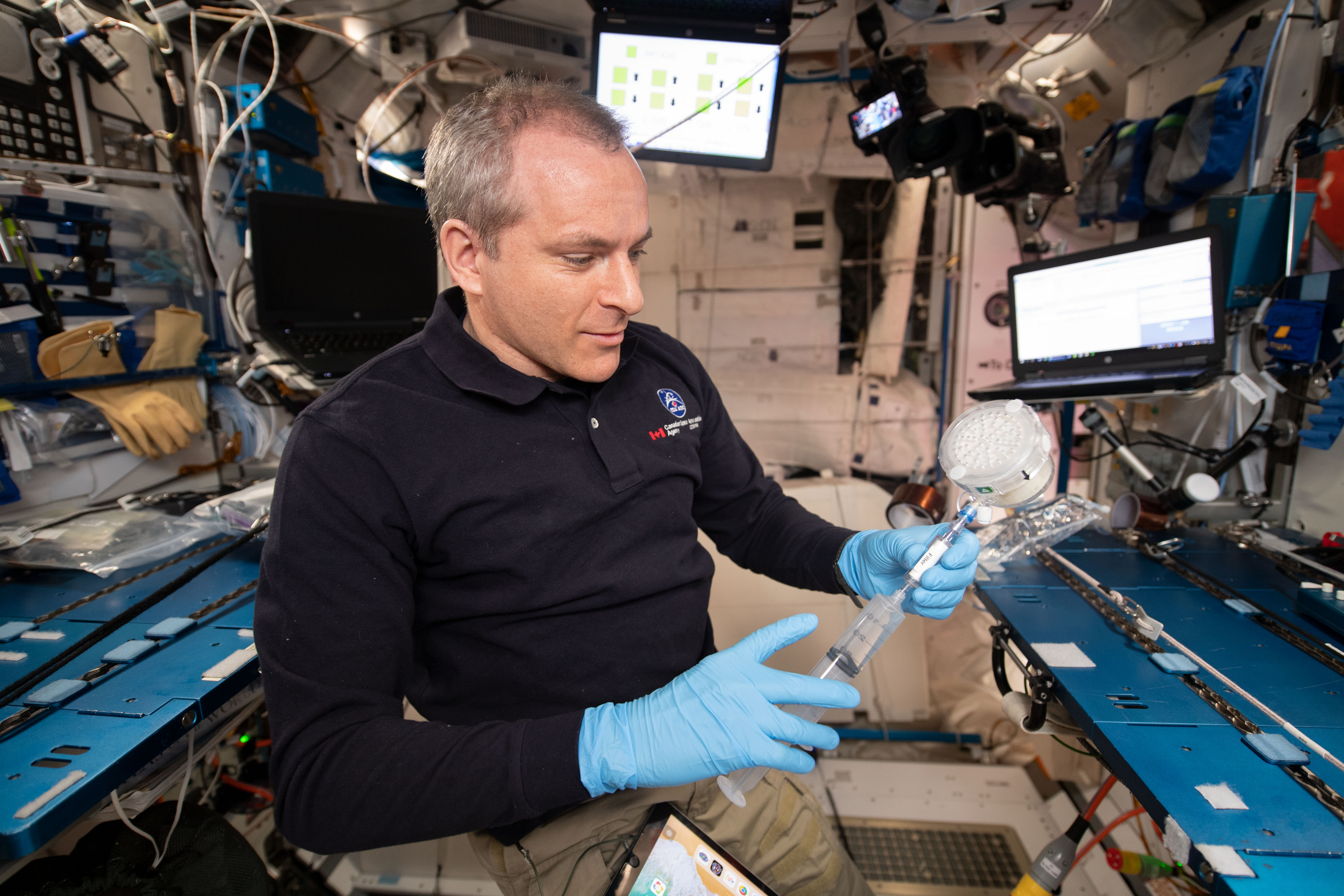 Astronaut David Saint-Jacques is seen performing an experiment on the space station. David is injecting liquid from a syringe into a small cylindrical container.