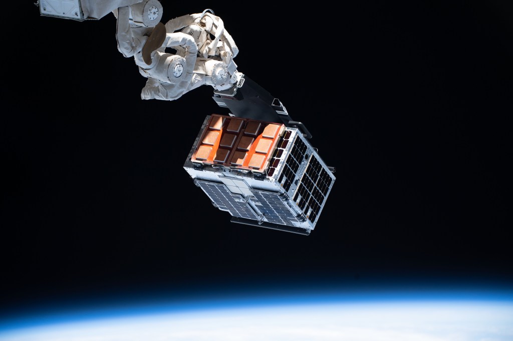An experiment is seen attached to a space station robotic arm. The experiment appears as a box with orange and blue colored solar panels on the sides. The Earth is seen out of focus at the bottom edge of the image.