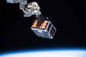 An experiment is seen attached to a space station robotic arm. The experiment appears as a box with orange and blue colored solar panels on the sides. The Earth is seen out of focus at the bottom edge of the image.
