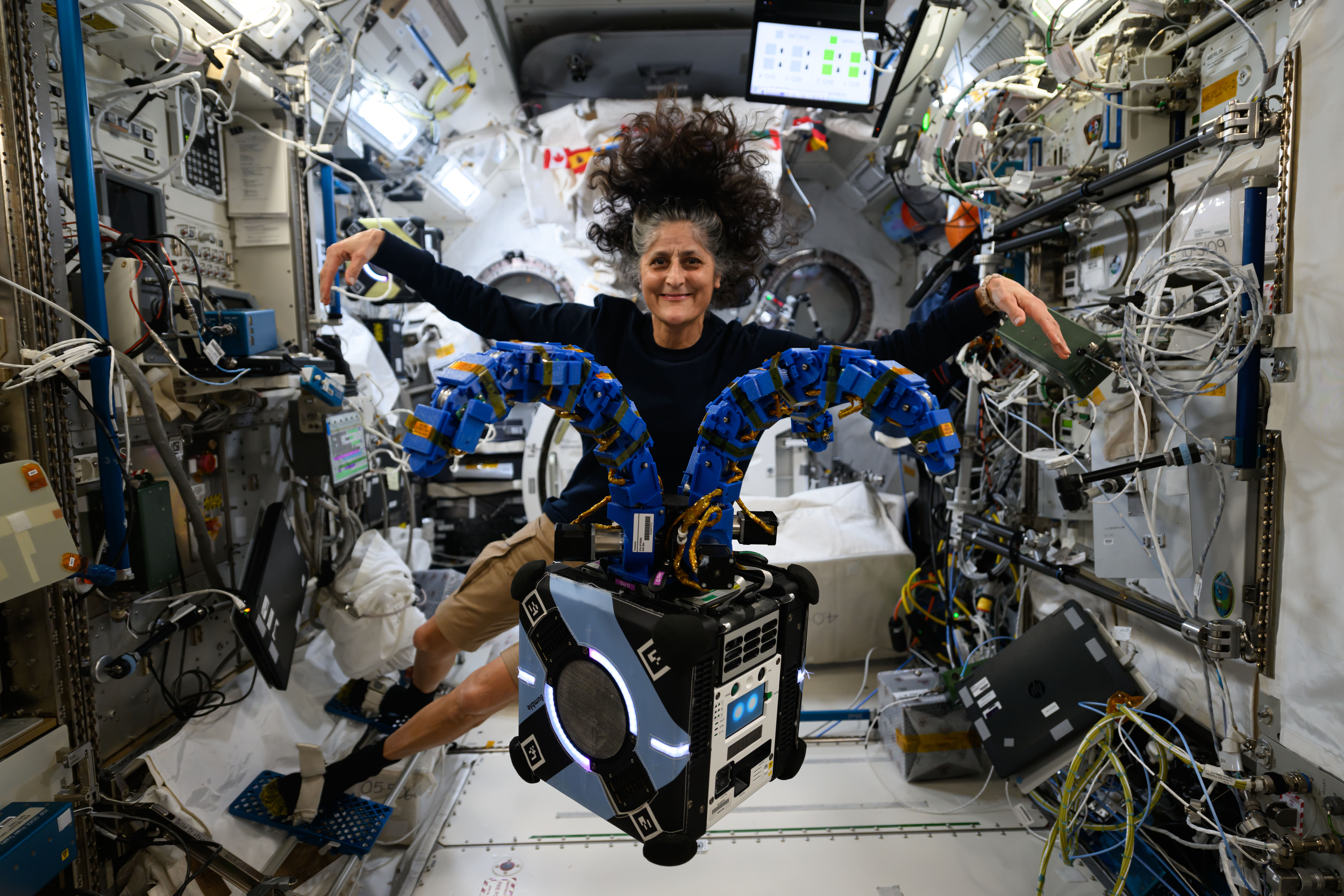 Astronaut Suni Williams poses with experiment hardware on the space station. The experiment hardware appears as a medium sized cube, with two blue arms at the top. The arms are protruding out from the top in arcs. Suni is floating behind the hardware, and she is holding her arms out, mimicking the experiment hardware.