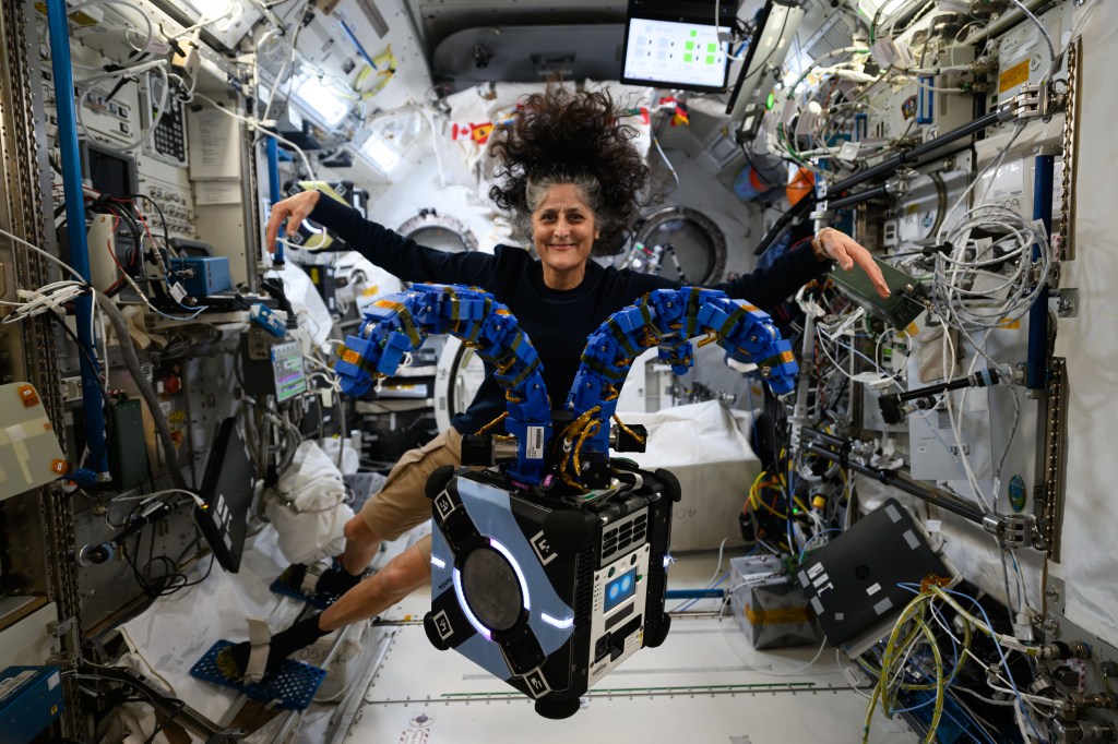 Astronaut Suni Williams poses with experiment hardware on the space station. The experiment hardware appears as a medium sized cube, with two blue arms at the top. The arms are protruding out from the top in arcs. Suni is floating behind the hardware, and she is holding her arms out, mimicking the experiment hardware.