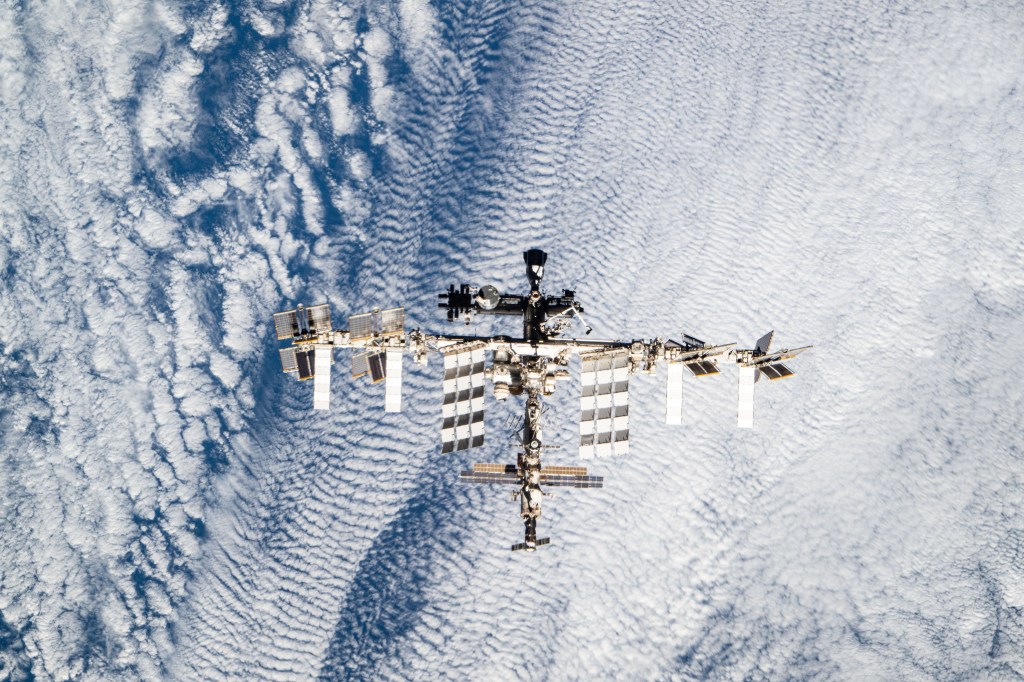 The International Space Station is seen orbiting over the Earth in this image taken by a crew member from another orbiting vehicle. The entire space station is seen at a distance, and the Earth appears in the background. The Earth appears as blue water with swirling white clouds.