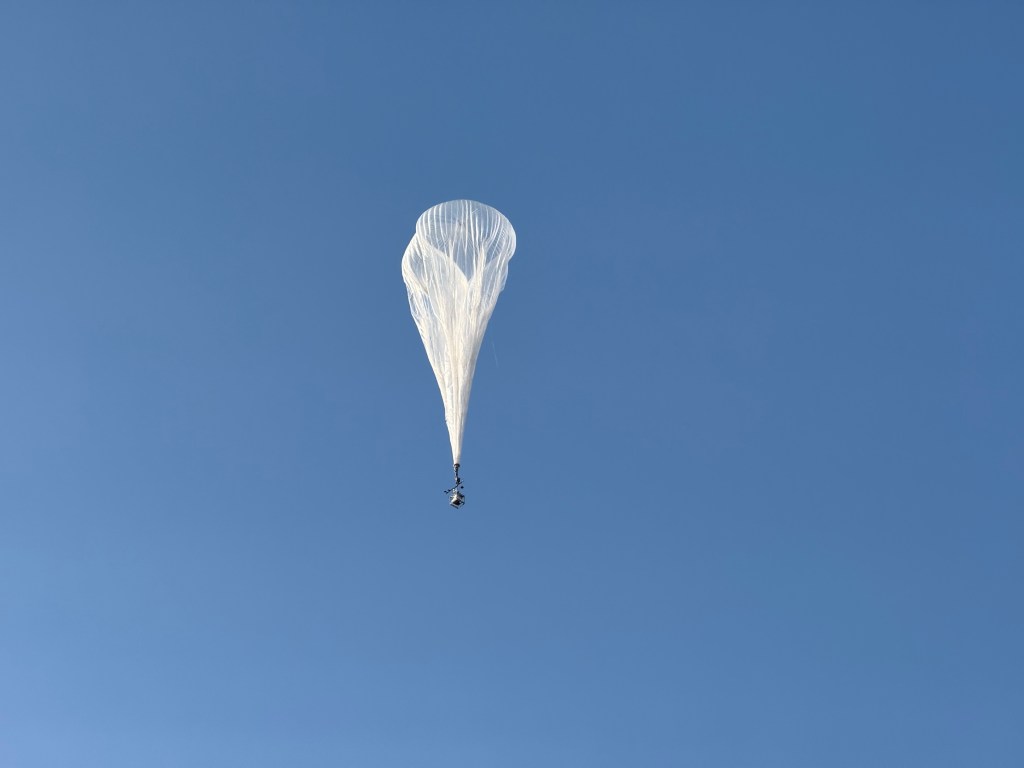High-altitude balloon with a square gondola hanging below floats in a bright blue sky.