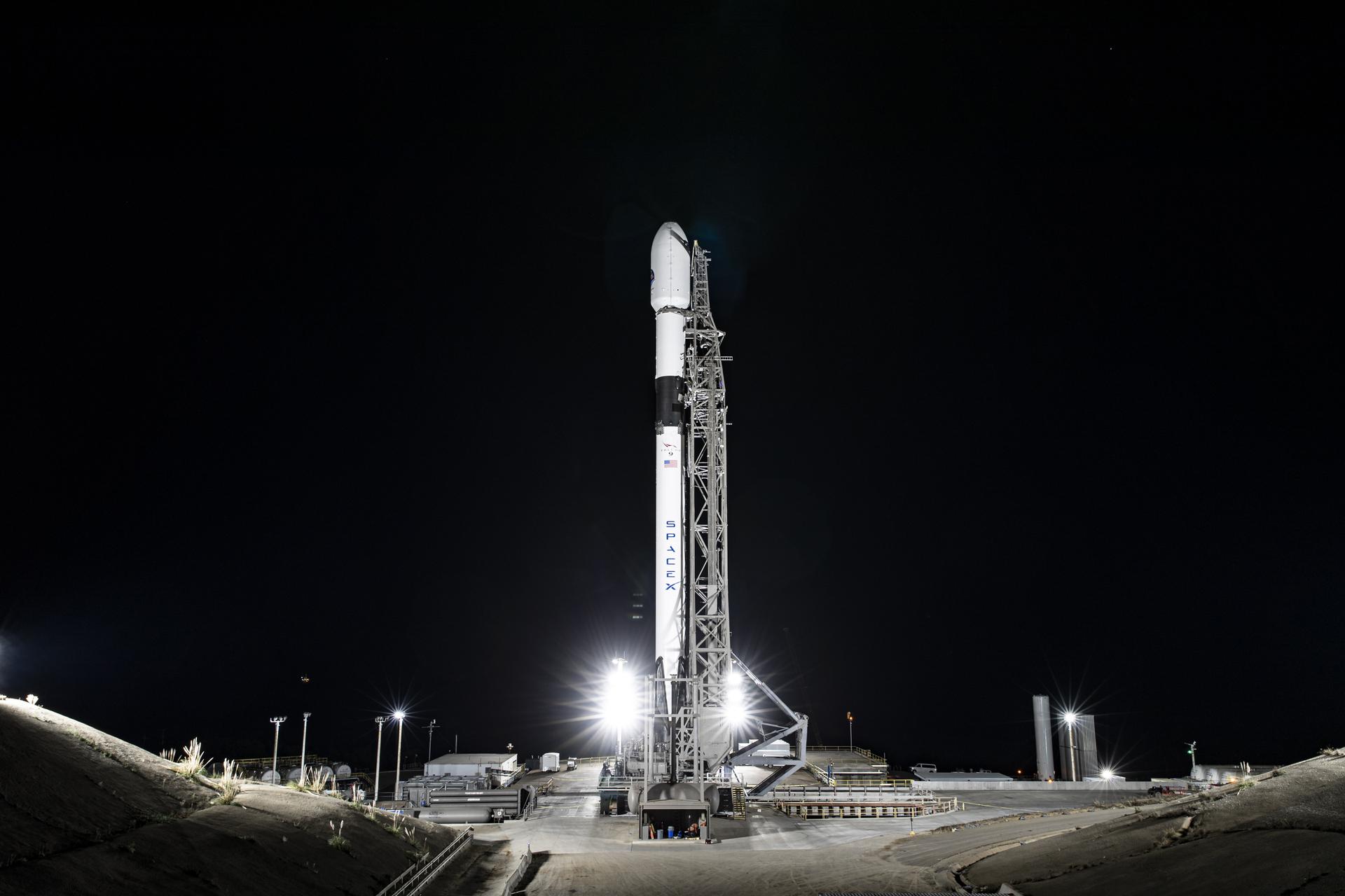 A wide-angle, distant photo of a white rocket on the launch pad, brightly lit, with a black night sky behind it.