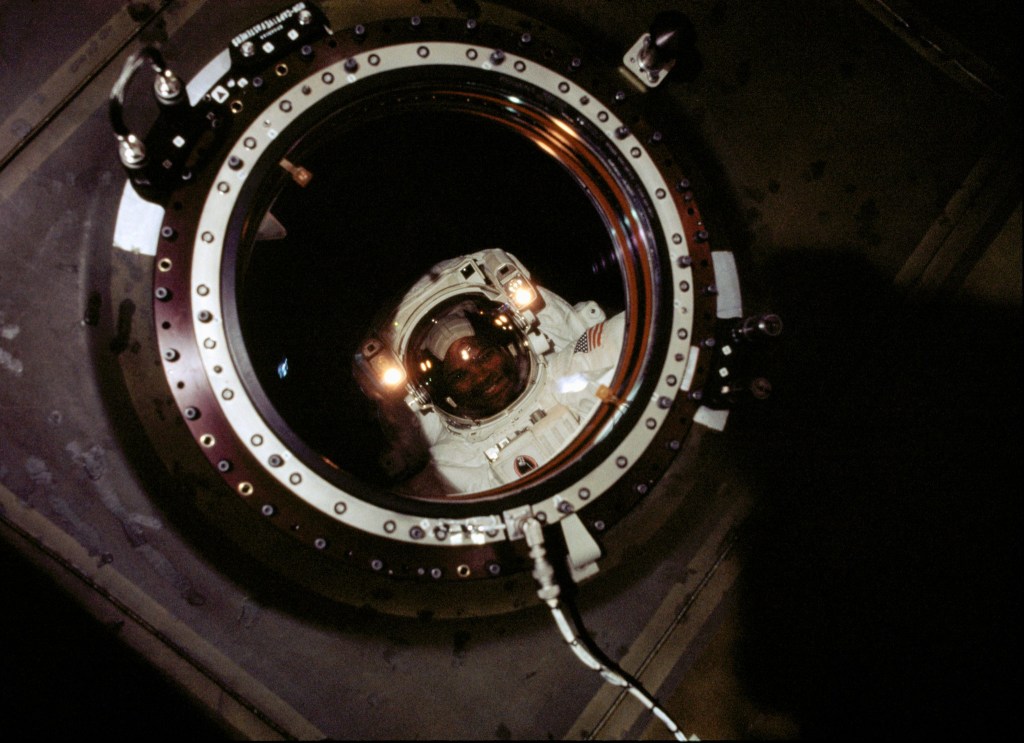 NASA astronaut Robert L. Curbeam, wearing a spacesuit, smiles as he looks through the lab window outside the Destiny module.