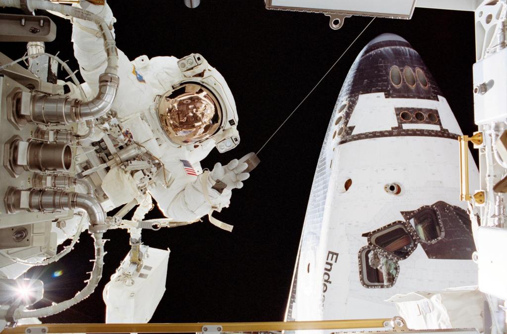 Astronaut John B. Herrington, in a space suit, waves from behind a portion of the space station. The forward section of the Space Shuttle Endeavor is to the right of Herrington.
