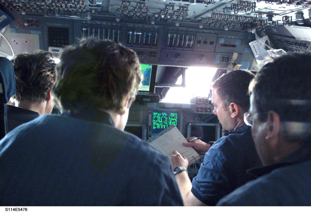 Viewing NASA astronauts, Eileen M. Collins, Wendy Lawrence, James M. Kelly and Charles J. Camarda from their backs, they sit at their stations in Space Shuttle Discovery. Kelly holds a clipboard with a pen.