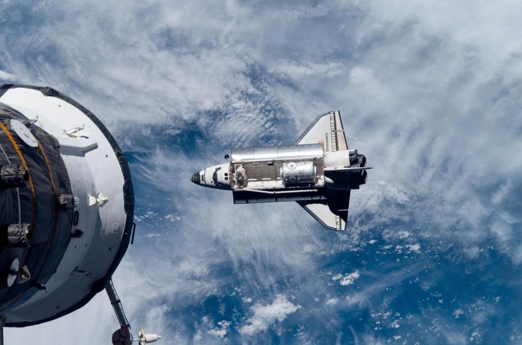 With a blue and white Earth in the background, Space Shuttle Discovery approaches a Russian spacecraft docked to the International Space Station on the left. The Harmony node is visible in Discovery's cargo bay.