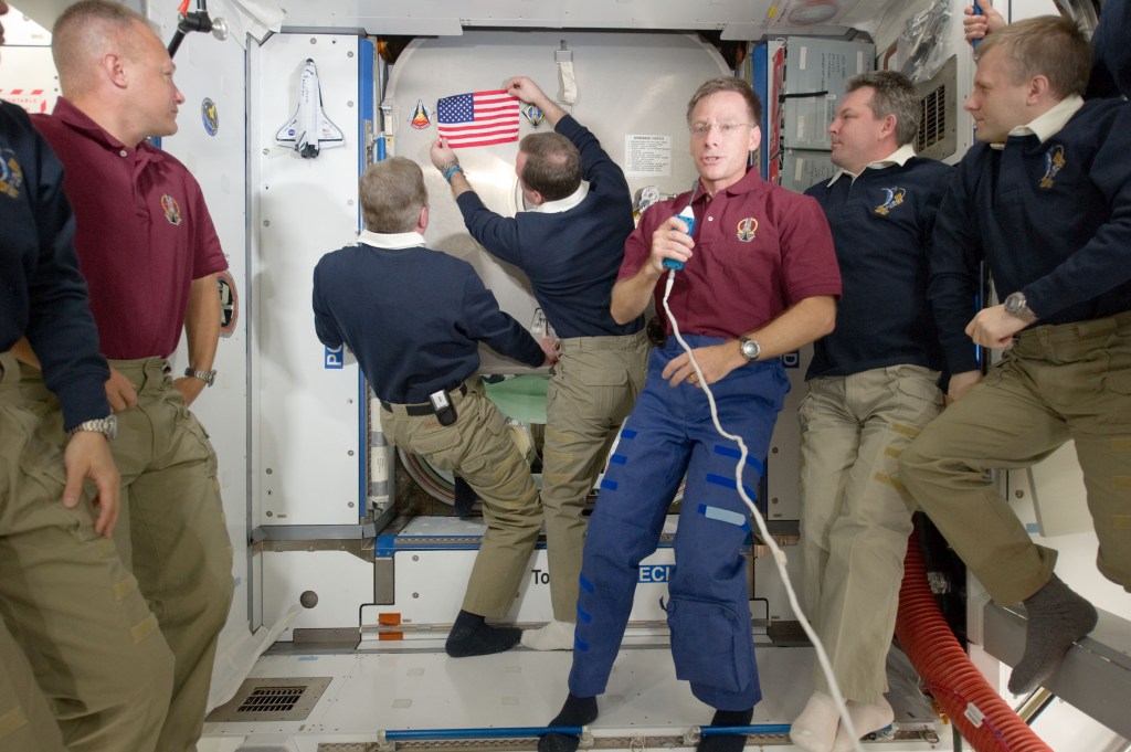 The Expedition 28 crew is in the background looking at the flag being placed on the Hatch of the Harmony module while Astronaut Christopher Ferguson holds a microphone looking at the camera.