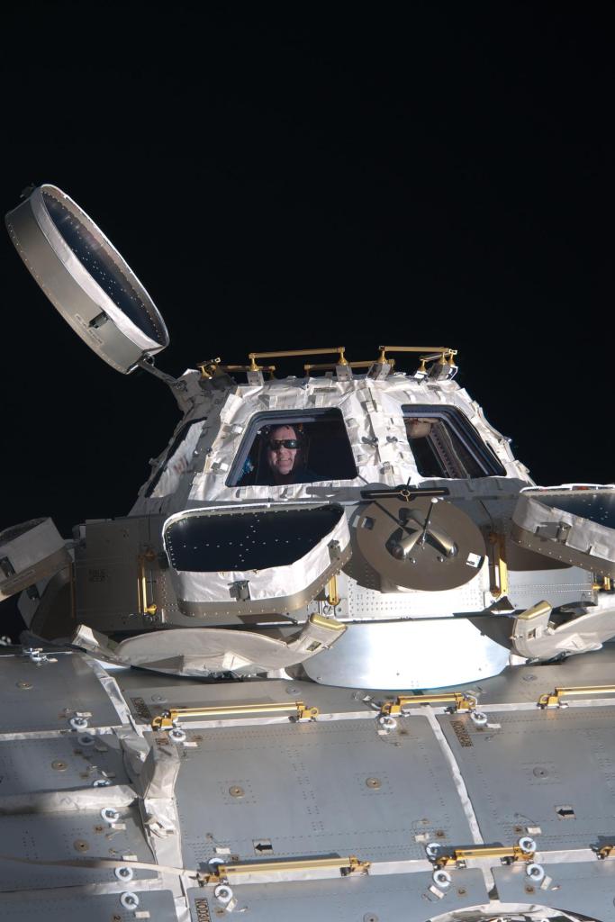 ESA astronaut Andre Kuipers looks through a window of the Cupola of the International Space station with an open hatch, backdropped by the blackness of space.