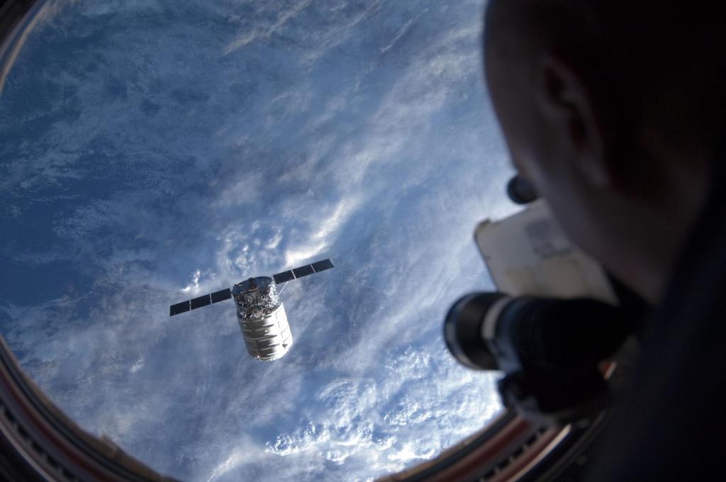 Through the window of the Cupola, ESA astronaut Luca Parmitano is off-frame to the right, holding an in-frame laser range finder, looking at Cygnus pictured on the left, backdropped by a blue and white Earth.