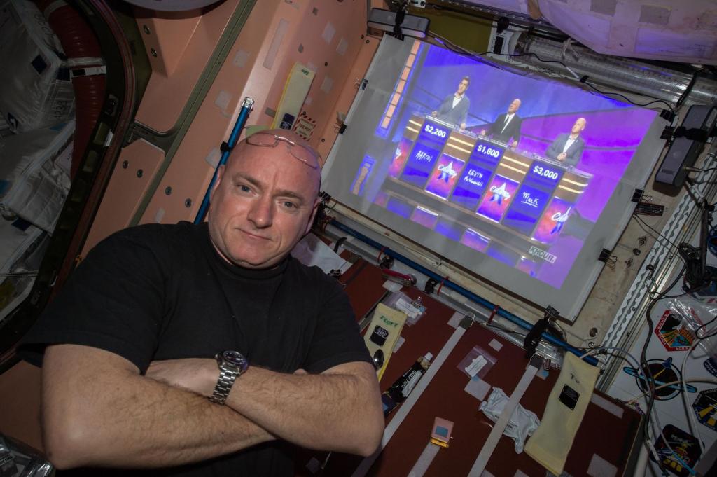 NASA astronaut Scott Kelly with a straight face, crosses his arms and is posed to the left of a screen showing a television show.