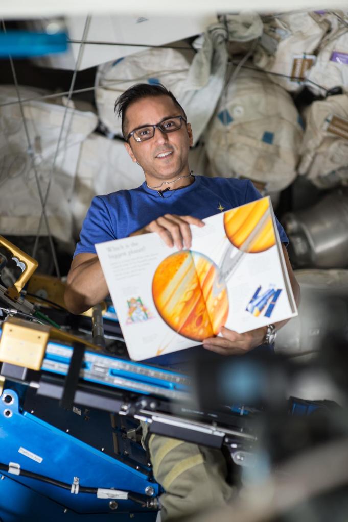 NASA astronaut Joe Acaba is smiling while he holds a children’s book with cargo in the background aboard the International Space Station.