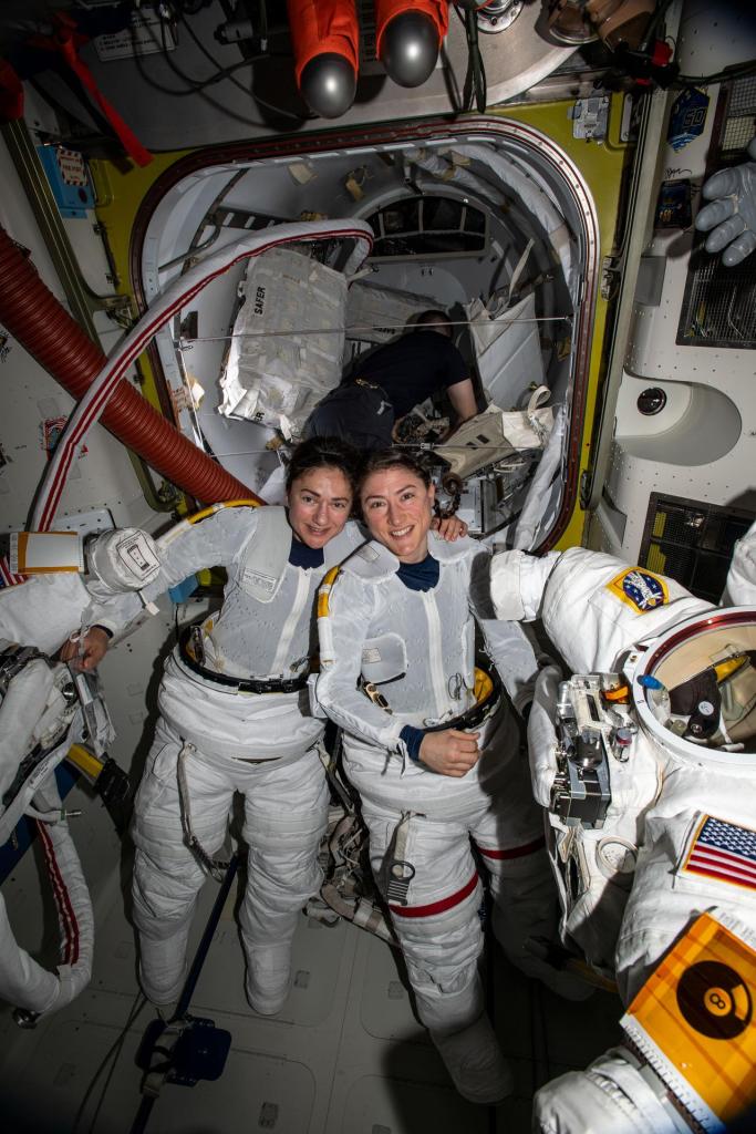 NASA astronauts Jessica Meir (left) and Christina Koch (right) are putting on their spacesuits while smiling at the camera.
