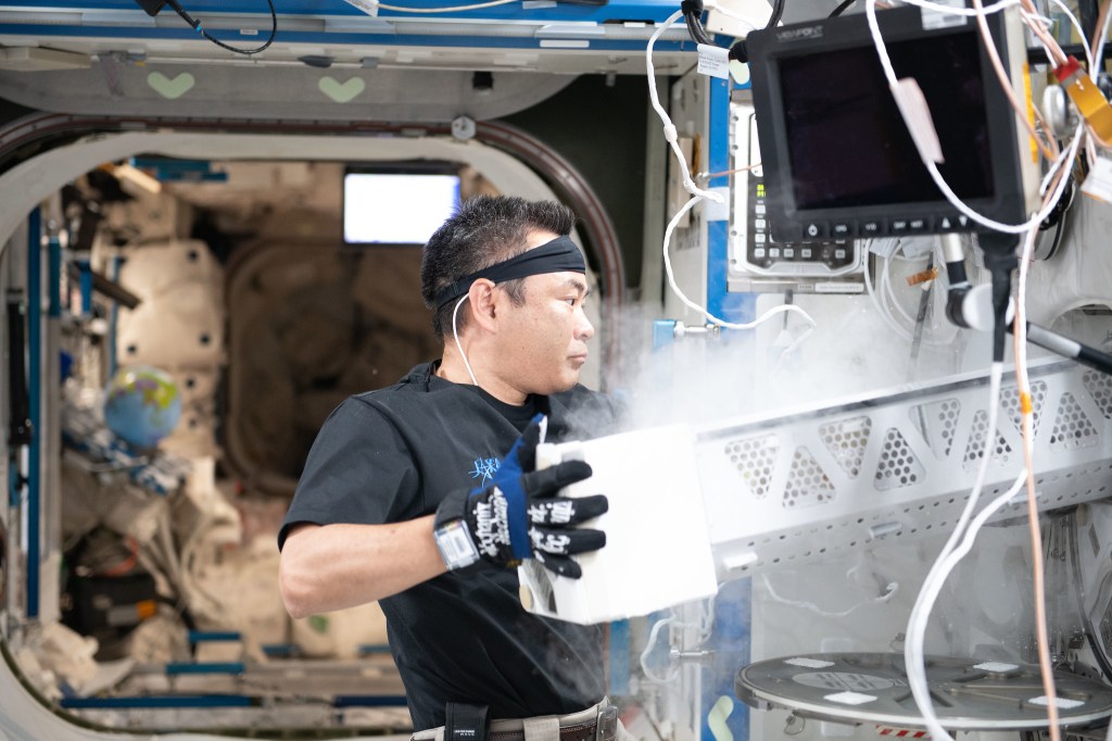 JAXA astronaut Akihiko Hoshide, wearing a black headband and gloves, stows samples inside a science freezer towards the right, surrounded by equipment and cables aboard the International Space Station.