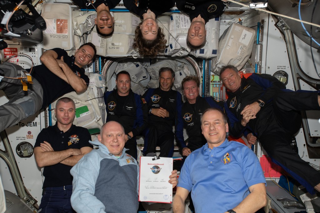 Eleven astronaut float together inside the International Space Station smiling for a group portrait. Three crew members are upside down at the top while others are right-side up, forming a tight cluster against the station’s interior wall.