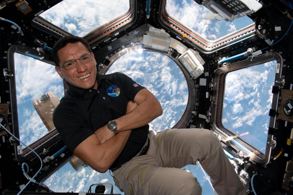NASA astronaut Frank Rubio smiles as he poses with his arms crossed inside the Cupola with a blue and white Earth in the background.