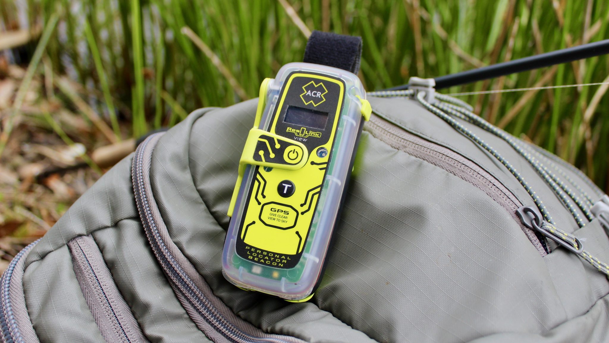 A close up picture of a green ACR PLB atop a bag in a forest setting
