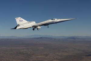 The X-59 quiet supersonic research aircraft flies above California. The plane has a distinctive shape with a long, sharply pointed nose. The nose is silver, while the rest of the body looks white. The words "NASA" and "X-59" are on the body of the aircraft. In the background, we can see brown earth below the plane as well as hazy mountains in the distance.